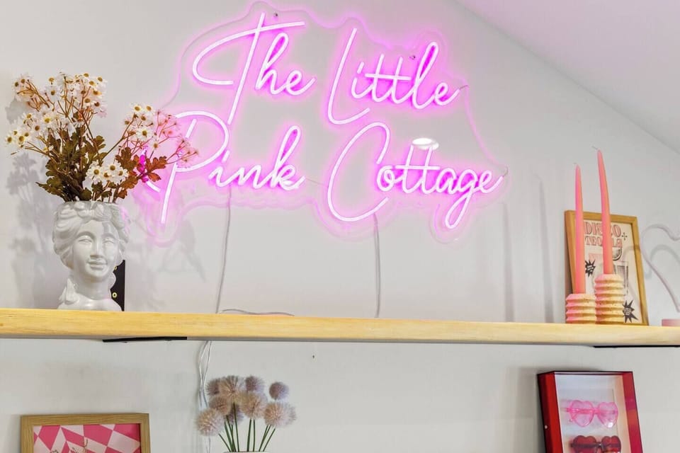 Welcome to "The Little Pink Cottage"! This charming room features a neon pink sign, a wooden shelf with a white bust vase, dried flowers, and pink candles, all set against crisp white walls. A perfect blend of cozy and stylish!