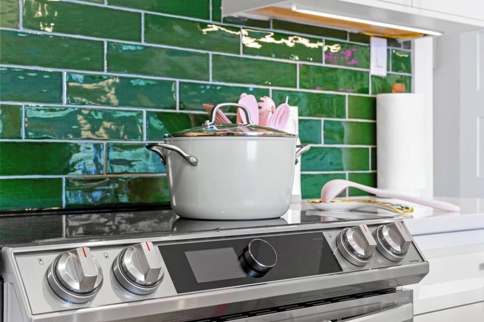 A modern kitchen featuring a stainless steel stovetop with a white pot, set against a glossy green subway tile backsplash. A white shelf above holds essentials like a paper towel roll and utensils, blending style with functionality.