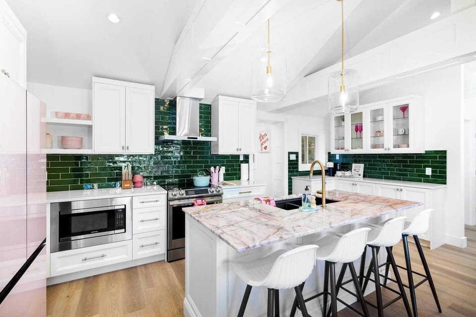 This bright, modern kitchen features white cabinets, a pink marble island, and vibrant green subway tiles. Stainless steel appliances, gold pendant lighting, and light brown hardwood flooring complete the stylish design.