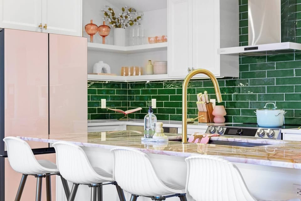 This vibrant kitchen features a pink fridge, white cabinetry, and a green subway tile backsplash. The marble island with a gold faucet is surrounded by white bar stools, while open shelves display warm, decorative accents.
