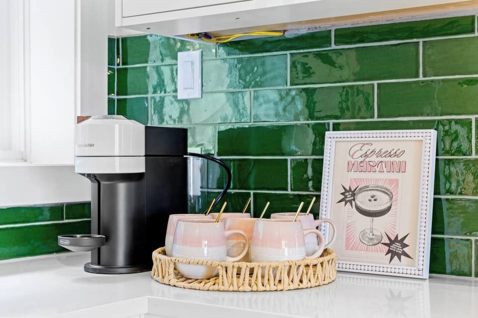 A charming kitchen corner featuring a green tile backsplash. The counter is adorned with a white and black coffee maker, pink ombre mugs on a woven tray, and a framed coffee-themed print with pink and green accents, perfect for your morning coffee.