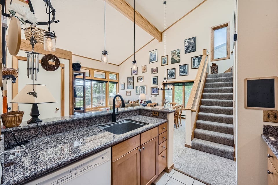 A kitchen with granite countertops, wooden cabinets, and a sink. Stairs lead upstairs on the right, and a wall decorated with framed pictures is visible in the background.