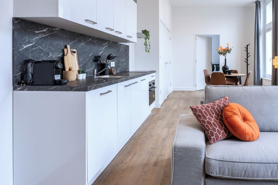 Modern living space with a sleek white kitchen, gray marble backsplash, cozy gray sofa accented with orange pillows, and a dining area featuring vibrant decor.