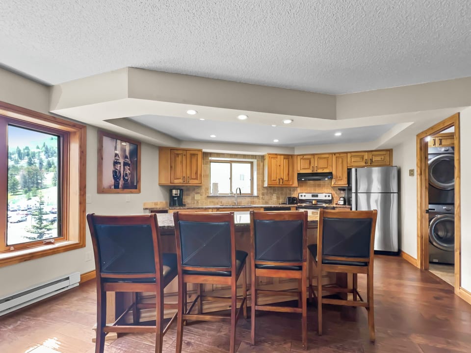 A modern kitchen features wooden cabinetry, a central island with stool seating, stainless steel appliances, and a window view of an outdoor landscape. A laundry area is visible on the right.