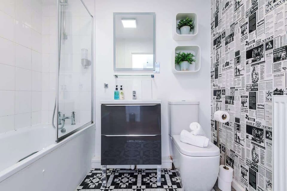 Modern bathroom with a sleek bathtub and glass shower screen, wall-mounted shower, and white tiled walls. Includes a dark vanity with mirror and shelf, stylish black-and-white flooring, and essential toiletries,creating a clean, contemporary space.
