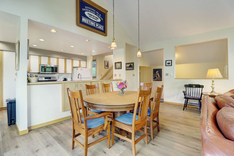 A brightly lit kitchen and dining area with wooden furniture, including a round dining table with six chairs, pendant lights above, and a sign reading "Meeting Place" on the wall separating the spaces.