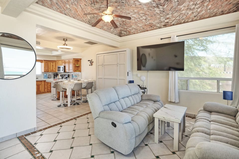 Spacious living room featuring plush reclining sofas, a large TV, and vaulted brick ceiling with ceiling fan—perfect for relaxing after a day at the beach.
