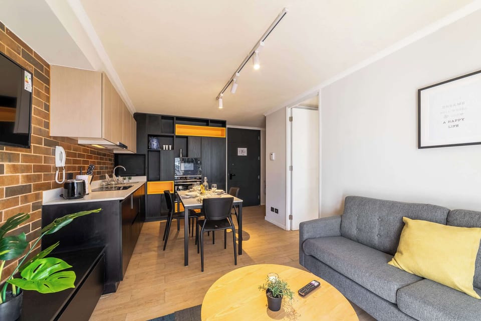 Modern open-plan living area with a fully equipped kitchen, dining table for two, and a cozy gray sofa with a mustard-yellow accent cushion.