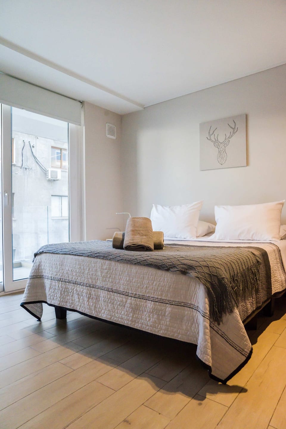 Bedroom with crisp white bedding, a textured gray quilt, geometric deer artwork, and a floor-to-ceiling window.