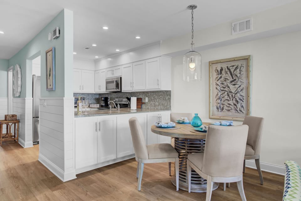 A modern kitchen with white cabinets, a small round dining table, beige chairs, and decorative elements like wall art and a pendant light.