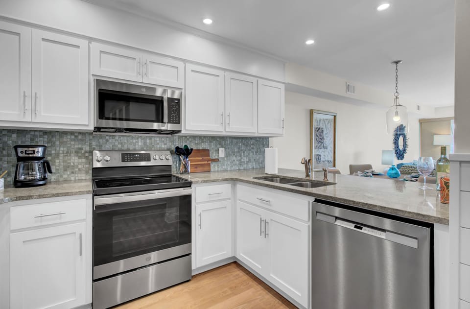 Modern kitchen with white cabinets, stainless steel appliances, electric stovetop, and double sink on a gray countertop. Bright lighting and hardwood floors complete the space.