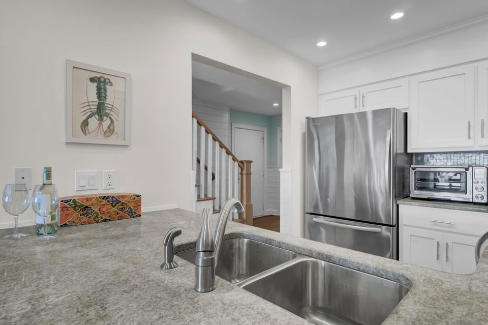 Modern kitchen with a stainless steel fridge, toaster oven, and double sink. A framed lobster print is on the wall. Stairs are visible through an open doorway.