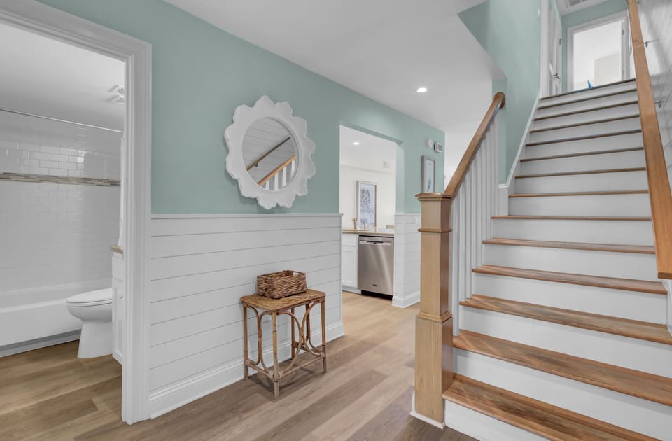 A hallway with light blue walls, wooden stairs, a round mirror, and a wicker basket on a small table. A bathroom and kitchen are visible.