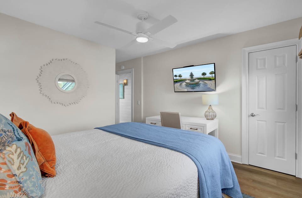 Bedroom interior with a bed, blue blanket, and decorative pillows. Wall-mounted TV above a small desk, round mirror on the wall, white door, and ceiling fan visible.