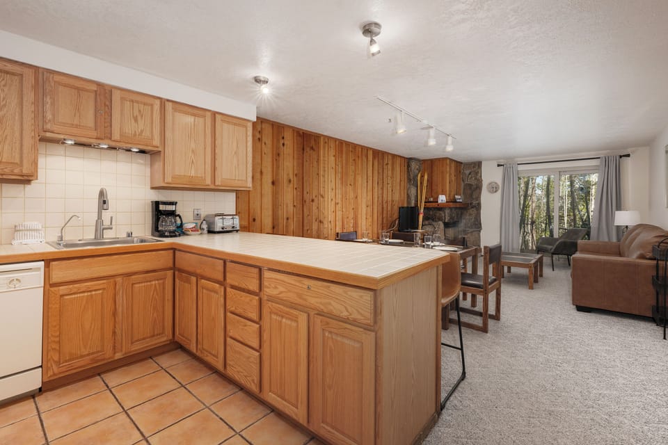 Your open-concept kitchen flows seamlessly into the cozy living area, where warm wood tones and a crackling fireplace create the perfect mountain retreat atmosphere.