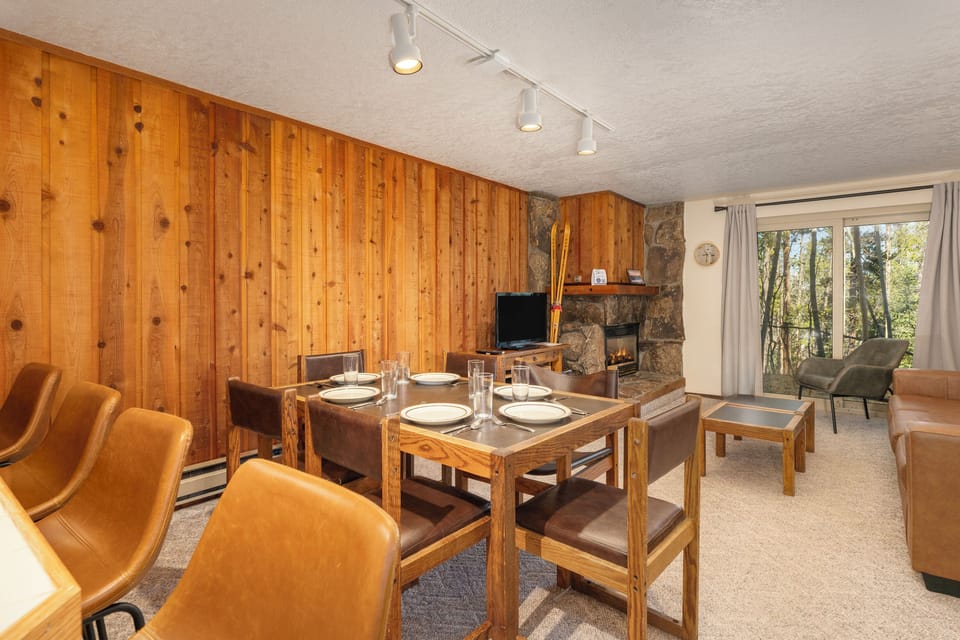 Gather around your dining table where warm wood walls and crackling fireplace create the perfect mountain retreat atmosphere.