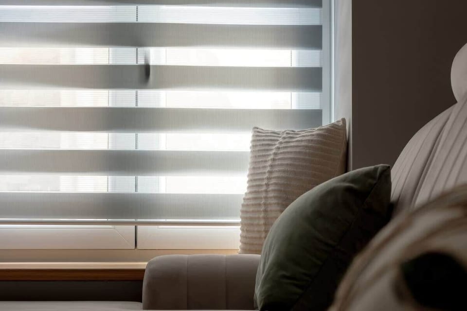 A close-up of a sofa next to a window with layered blinds, featuring soft lighting and textured fabrics.