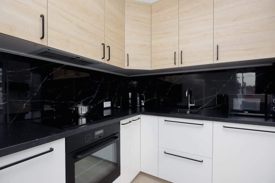 A sleek, L-shaped kitchen with light wood cabinetry, black glossy backsplash, and built-in appliances, designed for modern functionality.
