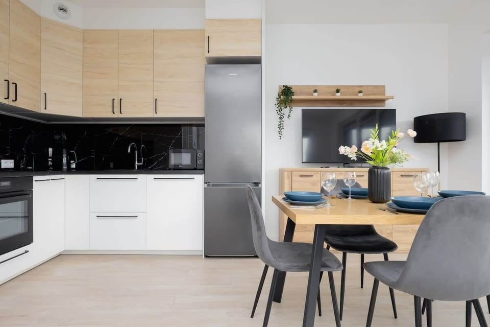 A sleek, L-shaped kitchen with light wood cabinetry, black glossy backsplash, and built-in appliances, designed for modern functionality.