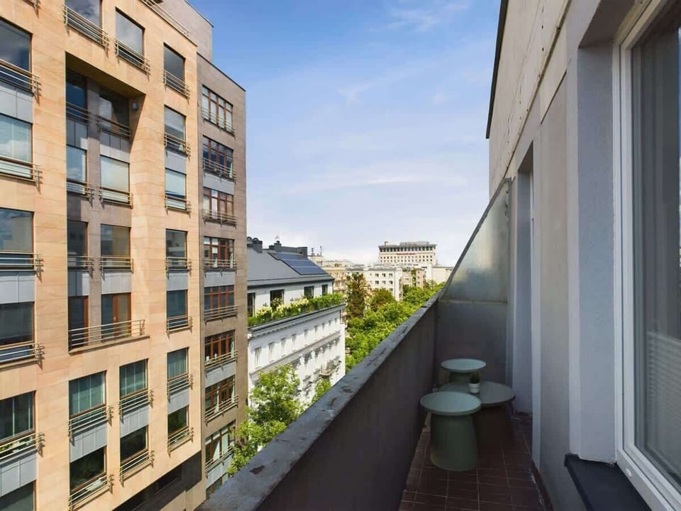 Balcony view overlooking modern residential buildings, blue sky, and tree-lined street.