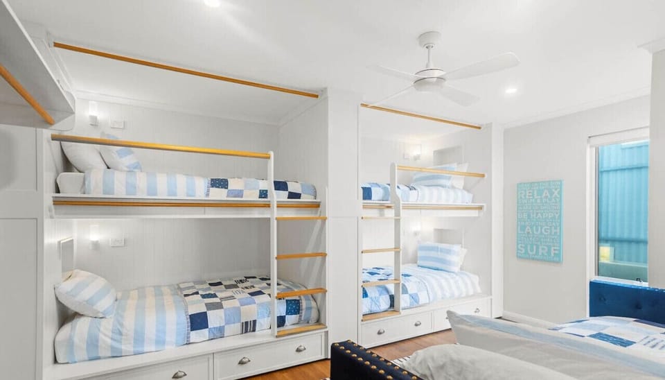 A cozy bunk bed room with white and blue striped bedding, featuring multiple bunk beds with wooden rails. The room has a clean, airy feel with a ceiling fan, and there is a motivational sign on the wall that reads `RELAX BE HAPPY LAUGH SURF.` The overall 