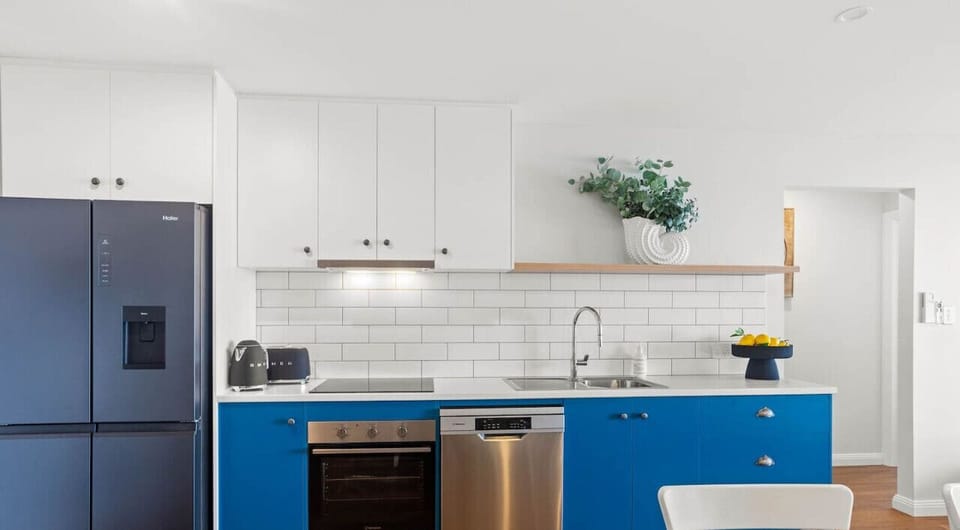 The kitchen includes a dark refrigerator, an oven, a dishwasher, and a countertop with a sink. There is also a decorative plant on a shelf above the countertop, adding a touch of greenery to the space. The overall look is clean and contemporary, with a wo