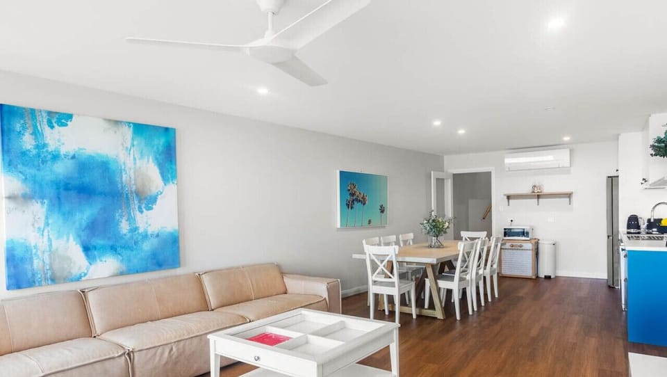 A stylish and modern living space with a beige sofa, a large blue abstract painting on the wall, and a wooden floor. The area also includes a dining table with white chairs, a kitchen with a blue cabinet, and various decorative elements, creating a cozy a