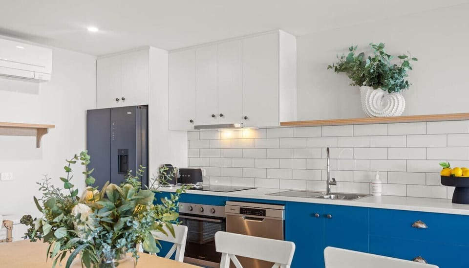 A modern kitchen with white cabinets and blue lower cabinets, featuring a stainless steel appliance suite, a vase of greenery on the table, and a decorative plant on a shelf above the tiled backsplash.