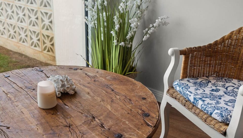 A rustic wooden round table with a candle and a decorative piece on it, accompanied by a wicker chair with a blue and white patterned cushion. The setting includes a tall plant with white flowers in the background and a decorative wall element, creating a