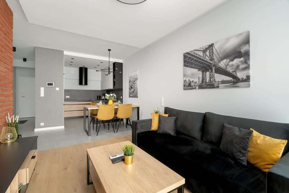 A bright open-plan living space with a gray couch, dining table, and small kitchen corner. A large black-and-white print decorates the wall.