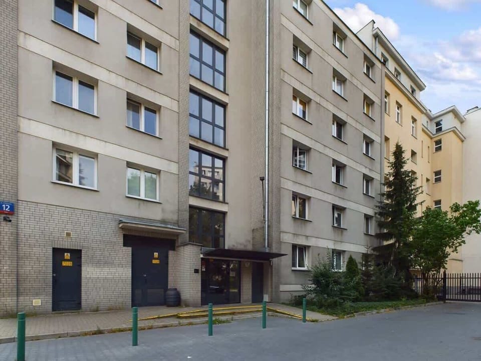 A view of the exterior of a modern apartment building with beige and brick detailing. The street is quiet and lined with trees.
