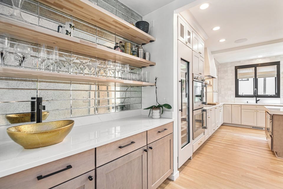 - Sip and serve from this stylish bar area featuring mirrored backsplash and floating shelves
- Display your glassware in style above sleek cabinets with soft wood tones and black pulls
- Gold bowl and potted orchid complete the refined look
