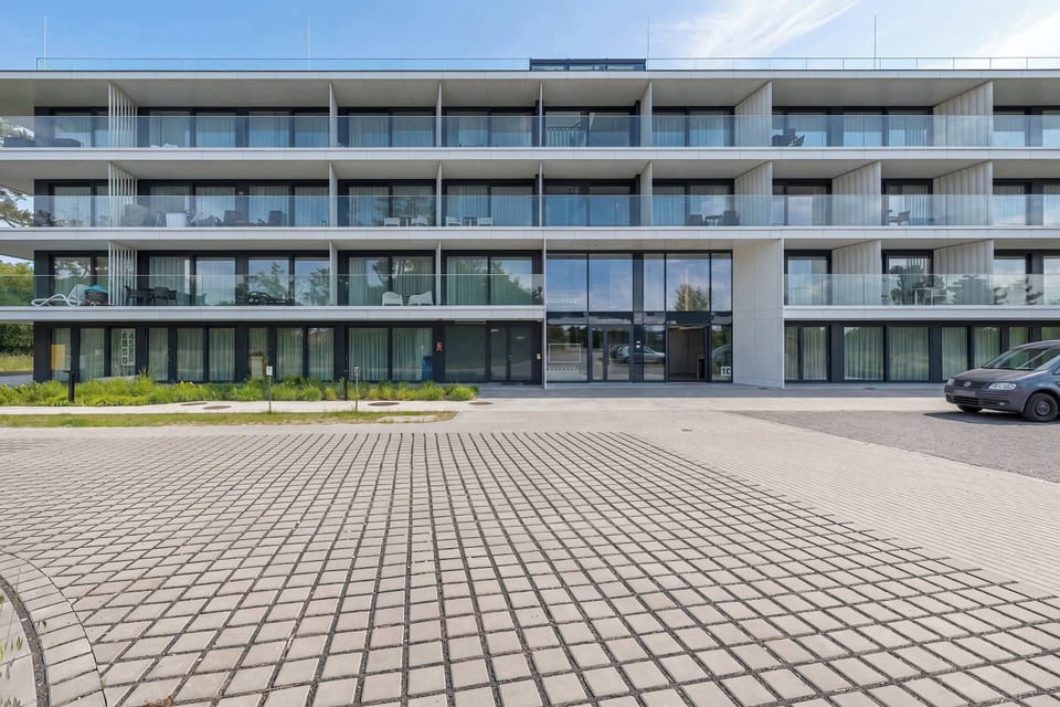 
A wide exterior view captures the entire facade of the modern residential complex featuring clean white lines and glass railings. A paved brick driveway sweeps past the front of the building under a clear blue sky.