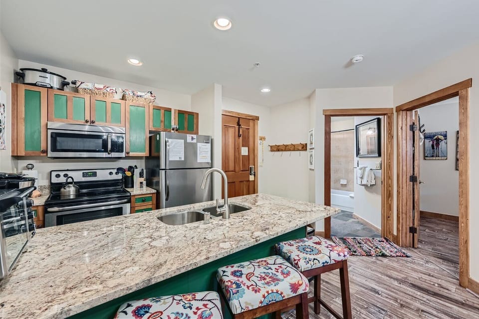 A modern kitchen with wood cabinets, granite countertops, stainless steel appliances, and a small island with three colorful barstools. An adjacent hallway leads to a bathroom.