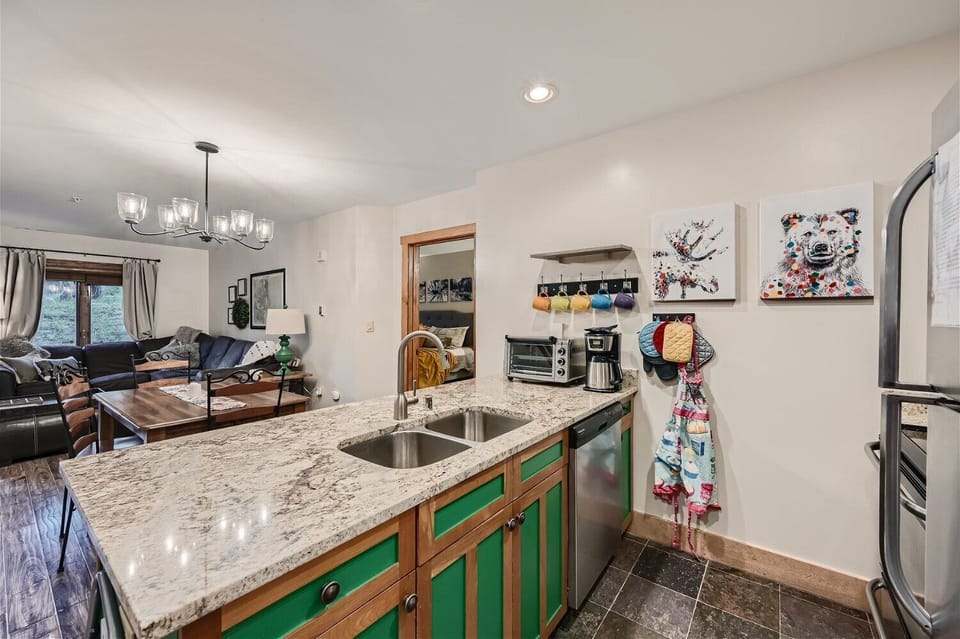 A kitchen with a granite countertop island featuring a double sink, green cabinets, and appliances. Art hangs on the wall beside coffee mugs and aprons. The living room is visible in the background.