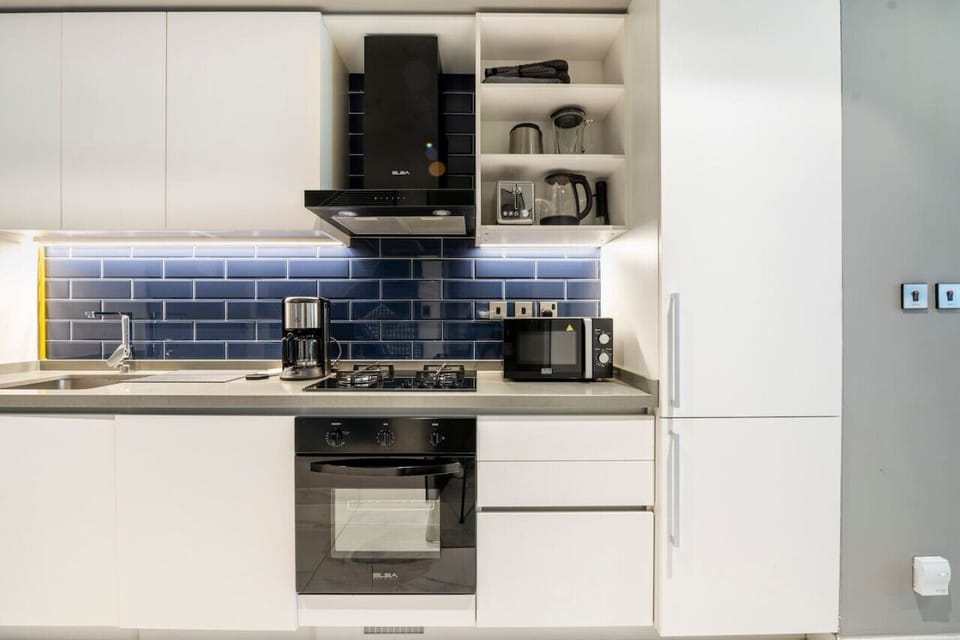 Modern, fully-equipped kitchen in a studio apartment at MAG 318, Business Bay. Features sleek white cabinets, blue backsplash, gas stove, oven, microwave, coffee maker, and essential appliances for a comfortable short-term rental in Dubai.