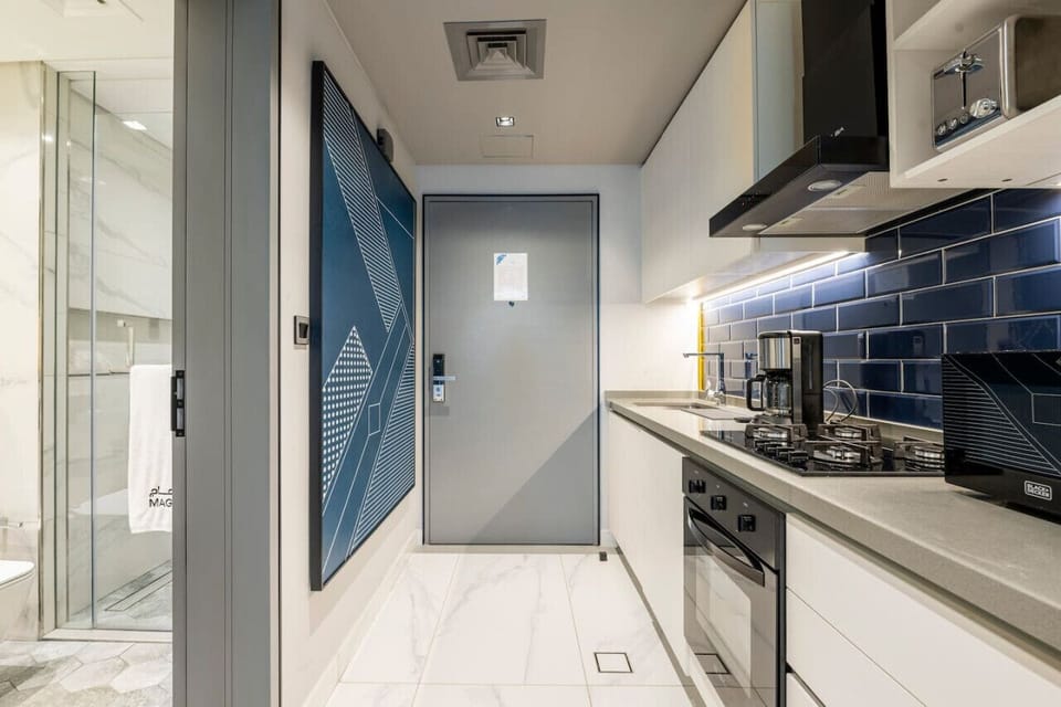 Entryway of a stylish studio apartment at MAG 318, Business Bay. Features a modern kitchen with blue backsplash, gas stove, and oven on one side and a sleek gray door opposite, making it an ideal holiday home in Dubai.