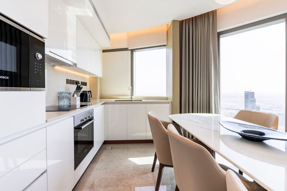 Modern kitchen and dining area in Address Harbour Point Tower 1, Dubai Creek Harbour. Features white cabinetry, integrated Bosch appliances, sleek countertops, and a white dining table with beige chairs. Large windows provide ample natural light.