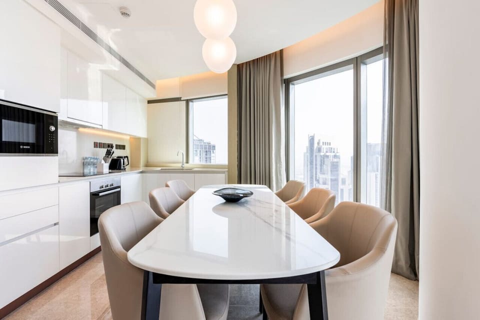 Elegant dining area in Address Harbour Point Tower 1, Dubai Creek Harbour. Features a sleek white dining table with beige chairs, modern pendant lighting, and large windows with city views. Adjacent modern kitchen with integrated appliances.