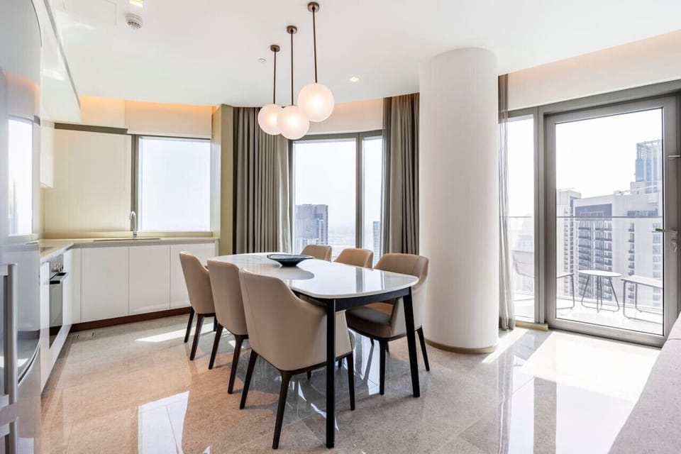 Bright dining area in Address Harbour Point Tower 1, Dubai Creek Harbour. Features a modern white dining table with beige chairs, elegant pendant lights, and large windows with city views. Adjacent to a sleek kitchen w/ white cabinetry and countertop