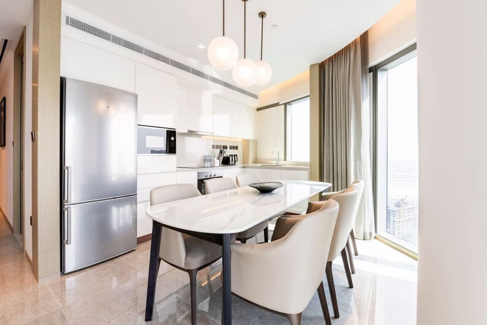 Stylish dining area in Address Harbour Point Tower 1, Creek Harbour. Features a sleek white dining table with beige chairs, modern pendant lights, and floor-to-ceiling windows. Adjacent kitchen boasts white cabinetry and stainless steel appliances.