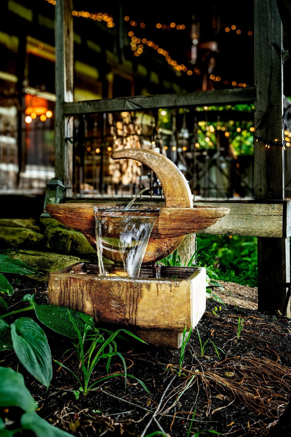 Rustic outdoor water fountain with flowing stream set against weathered wood porch and surrounding greenery.