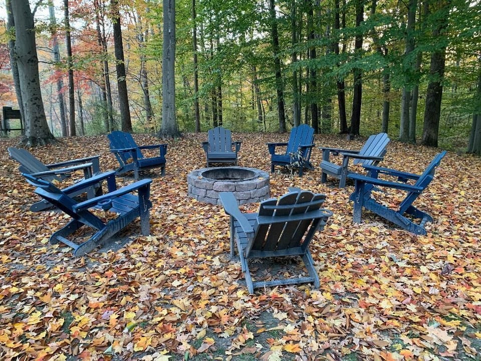 Circle of blue Adirondack chairs surrounds a stone fire pit on a ground covered with colorful autumn leaves.