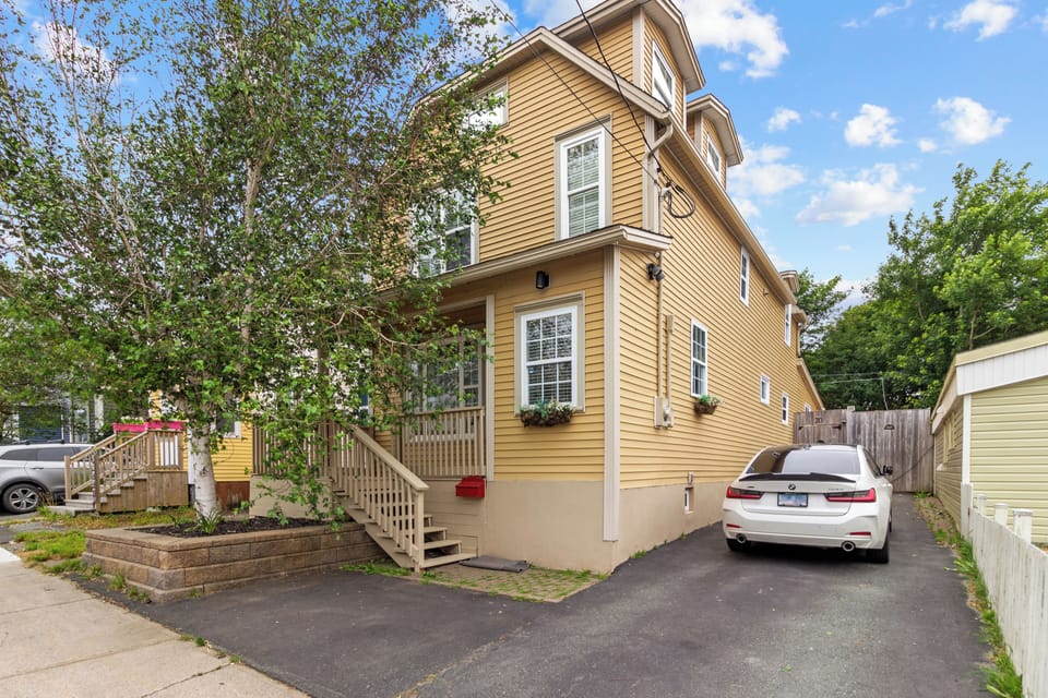 The front exterior of the house features a well-maintained yard, a charming porch, and a driveway with ample parking space. The yellow siding and large windows create a welcoming facade.

