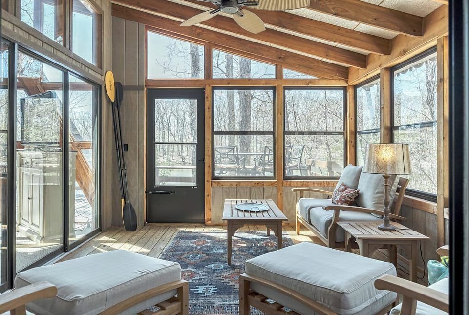 Furnished screened porch with cushioned wood-frame seating, ottomans, coffee table, side table, ceiling fan and broad wooded views.