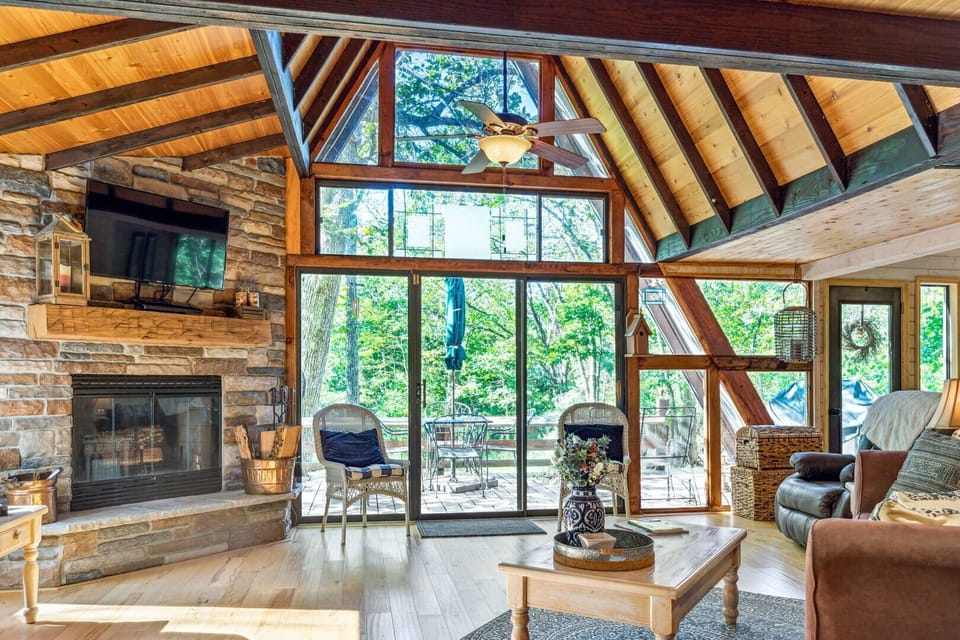 Vaulted living room with stone fireplace, TV, seating, and wall-height windows opening toward the deck and woods.