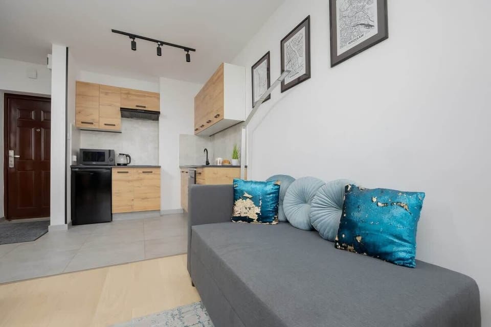 A compact yet inviting living room with a grey couch and vibrant teal pillows. Great use of space with direct access to the kitchen.