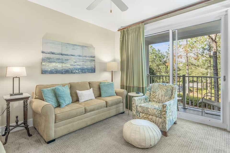 Bright coastal living room with plush seating, soothing decor, and sliding doors opening to a private balcony with leafy views.