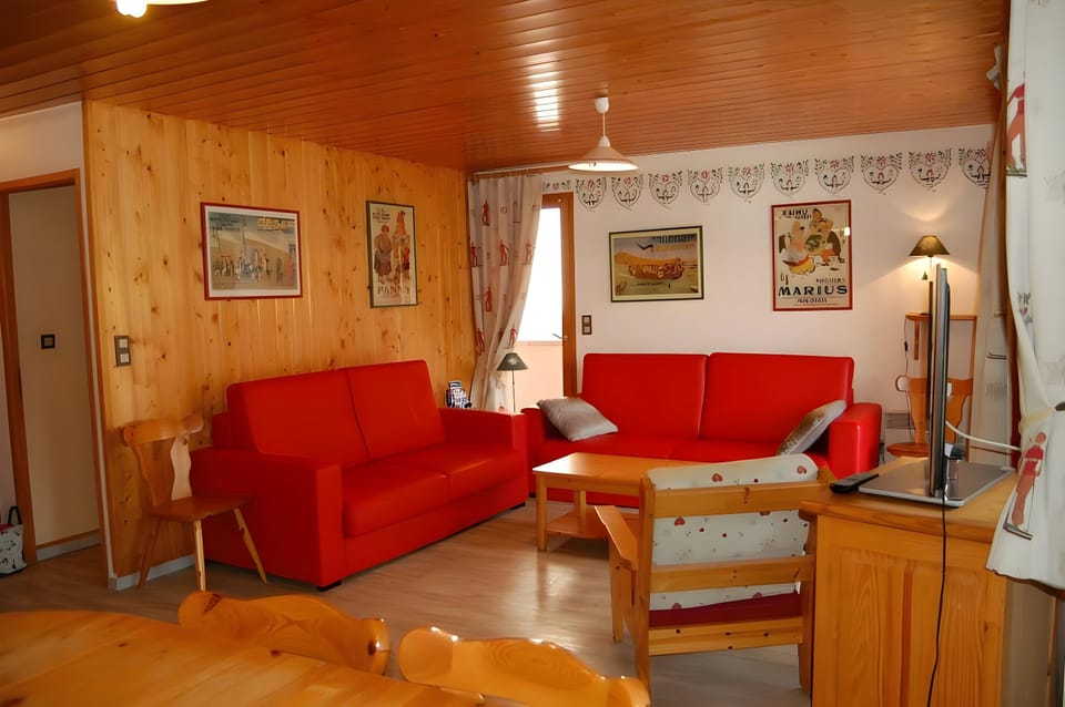 The spacious living area is designed for comfort and relaxation, featuring two red sofas, a wooden coffee table, and a charming armchair with a floral pattern. The wooden floorboards create a warm ambiance, complemented by the wood-paneled walls showcasin