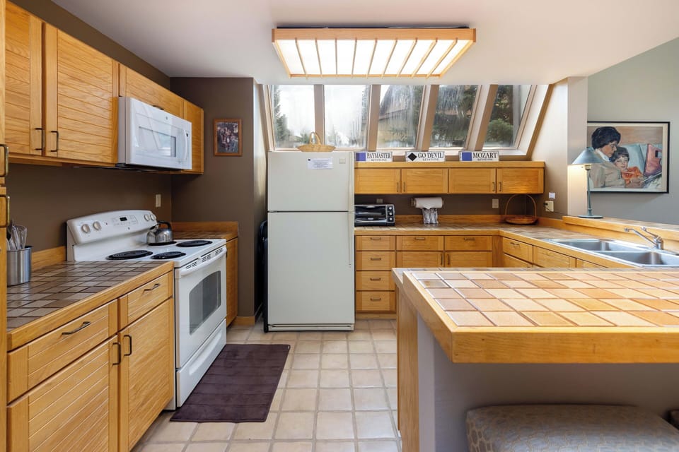 A kitchen with wooden cabinets, white appliances including a stove, microwave, and refrigerator, and a tile countertop. Natural light enters through angled windows above the sink.